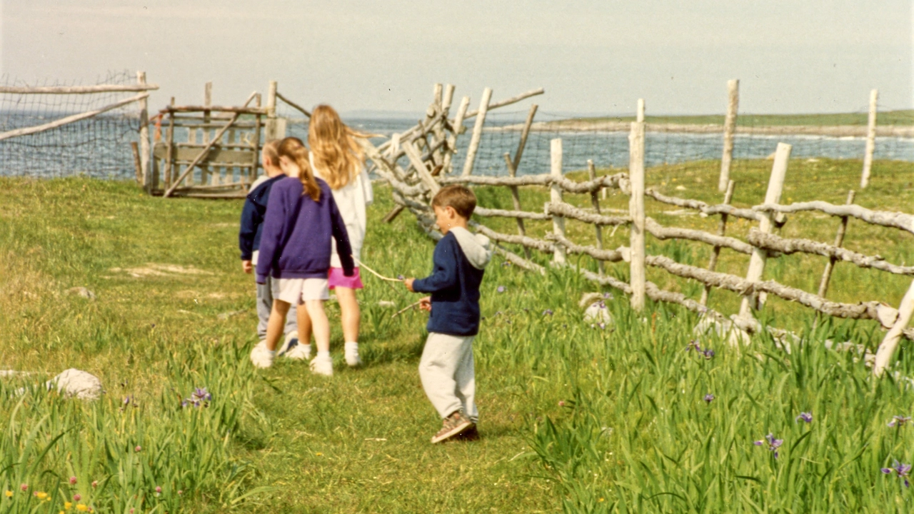 Nolan Franklin as a child in a blue hoodie walking with his siblings and cousins along a grassy path toward the beach in Cape Breton, with wildflowers, irises, a wooden fence, and the ocean beyond.