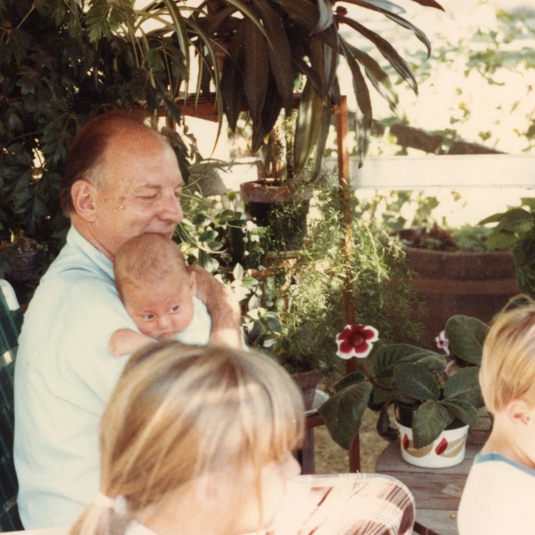 Nolan Franklin as a baby being held by his grandpa in a plant-filled gazebo, with his siblings in the foreground.