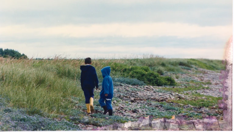 Two children walking along a rocky Cape Breton shoreline with beach grasses and low coastal plants.